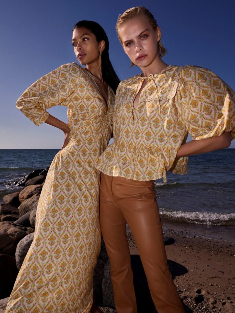 Două femei: una poartă o rochie lungă cu imprimeu și cealaltă o bluză cu imprimeu și mâneci bufante și pantaloni maro din piele