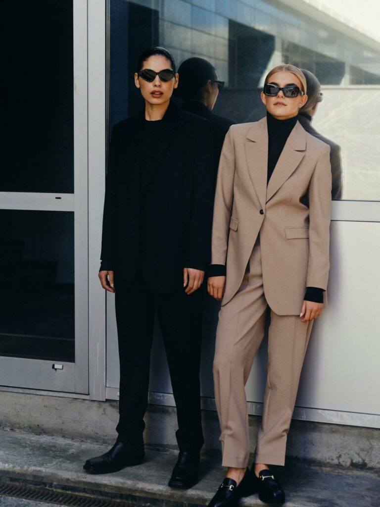 Două femei: una poartă un costum negru, ochelari de soare negri și cealaltă un costum bej, o bluză pe gât neagră și ochelari de soare negri