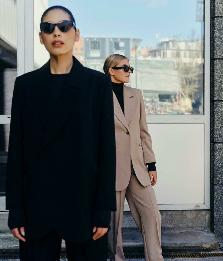 Două femei: una poartă un total look negru, ochelari de soare negri și cealaltă un costum bej, o bluză pe gât neagră și ochelari de soare negri