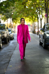 Nikki Huang poartă o cămașă cu guler înalt cu volane cu model de dantelă roz neon, un sacou lung roz neon, pantaloni cu picioare largi roz neon asortați, pantofi negri cu tocuri din piele lucioasă