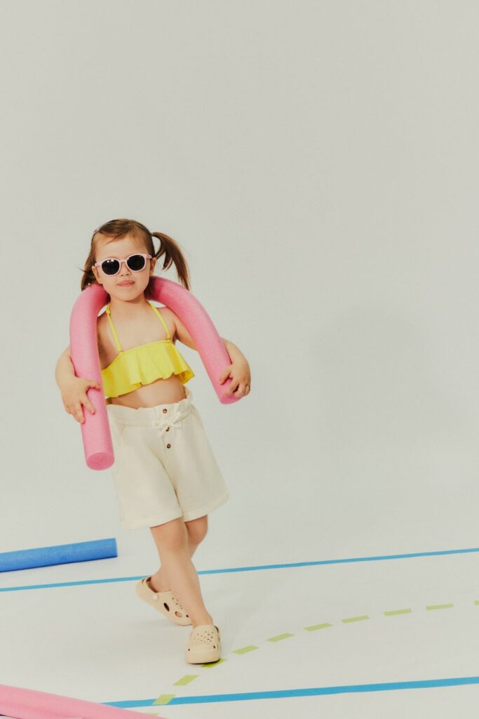 Fetiță cu top galben, pantaloni scurți albi, șlapi crocs și ochelari de soare