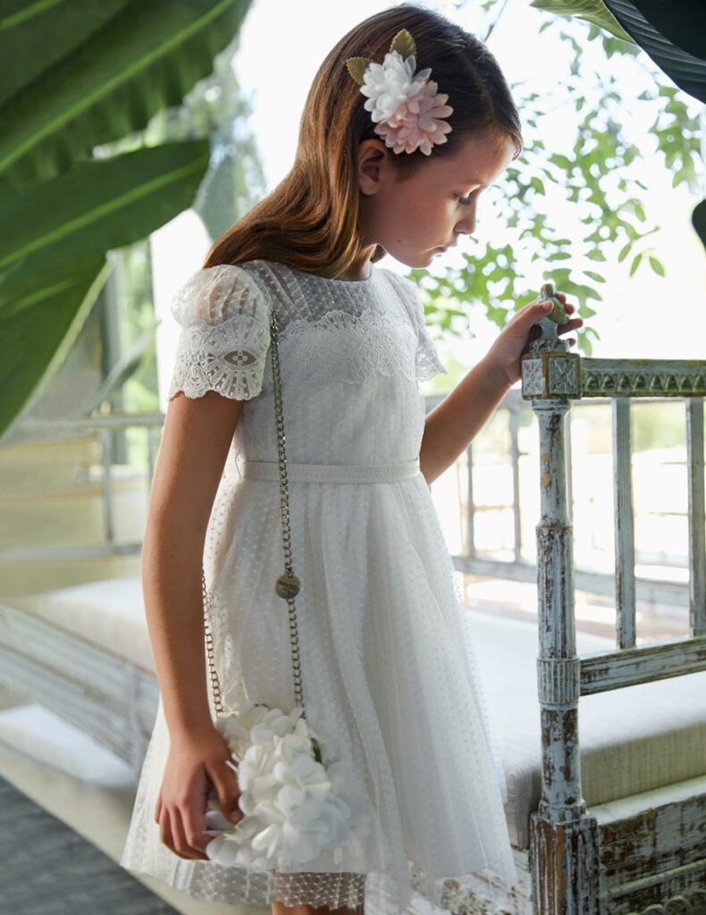 Fetiță cu o rochie albă din dantelă, o geantă albă tip flori și flori colorate în păr