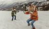 Doi copii se joacă în zăpadă îmbrăcați în geci groase de iarnă, bocanci, căciuli și fulare