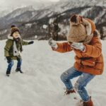 Doi copii se joacă în zăpadă îmbrăcați în geci groase de iarnă, bocanci, căciuli și fulare