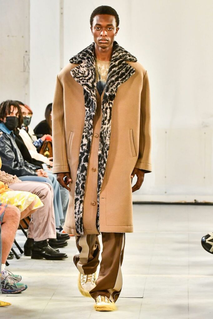 Model care poartă un palton lung bej cu gulere cu imprimeu animal,  pantaloni maro și bej sneakerși bej