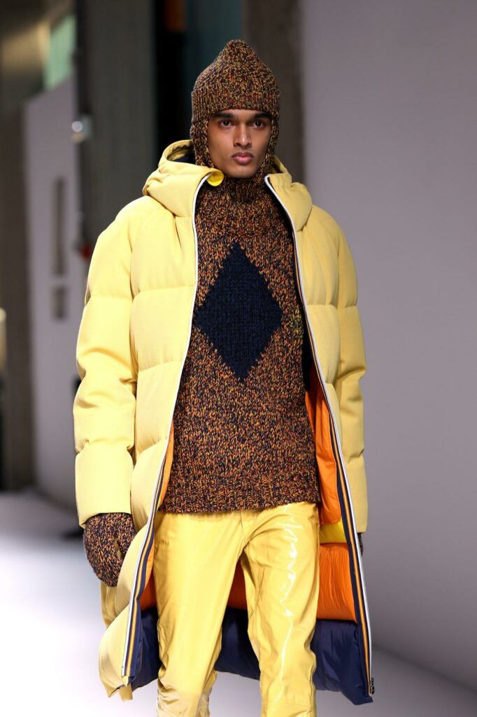 Model care poartă un palton lung galben din puf, pantaloni galbeni, pulover, căciulă și mănuși tricotate