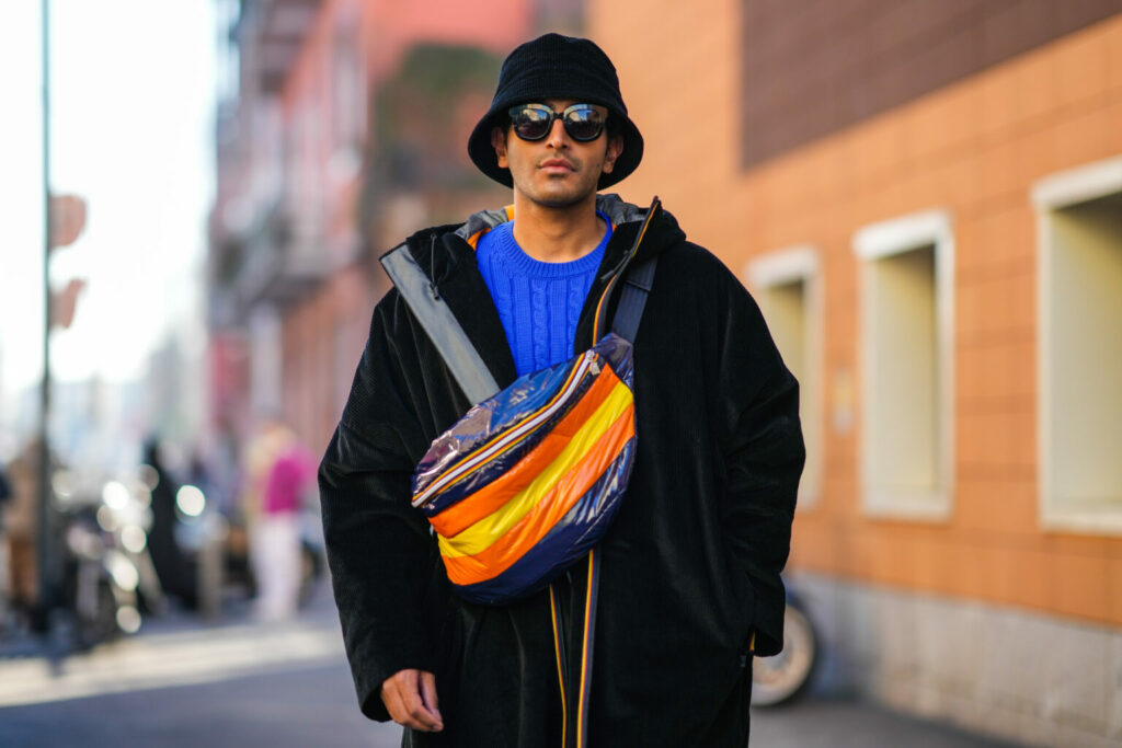 MILAN, ITALY - JANUARY 17: Rahi Chadda wears a black velvet bob / hat, black sunglasses, a royal blue electric braided wool pullover, a black hoodie oversized long zipper coat, a navy blue / orange / yellow oversized zipper fanny pack bag, outside the K-way fashion show, during the Milan Men's Fashion Week - Fall/Winter 2022/2023 on January 17, 2022 in Milan, Italy. (Photo by Edward Berthelot/Getty Images)