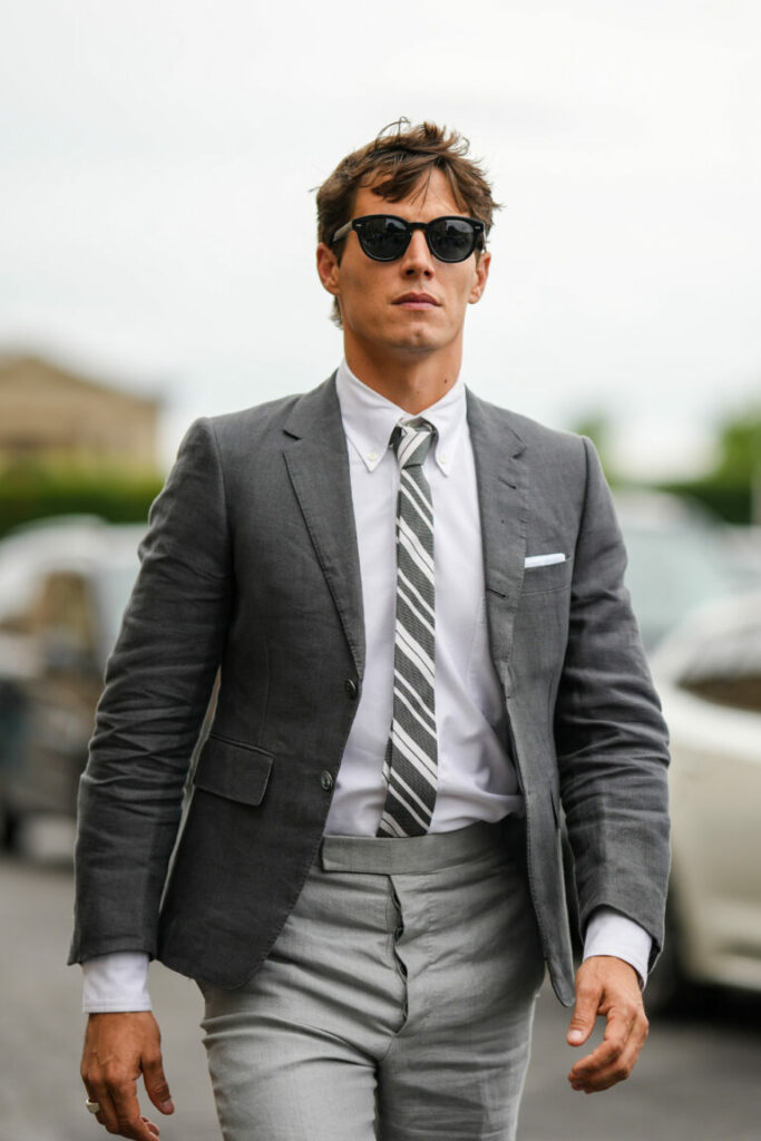 PARIS, FRANCE - JUNE 26: A guest wears black sunglasses, a white shirt, a gray and white striped print pattern tie, a dark gray blazer jacket, pale gray suit pants, rings, outside the Thom Browne show, during Paris Fashion Week - Menswear Spring/Summer 2023, on June 26, 2022 in Paris, France. (Photo by Edward Berthelot/Getty Images)