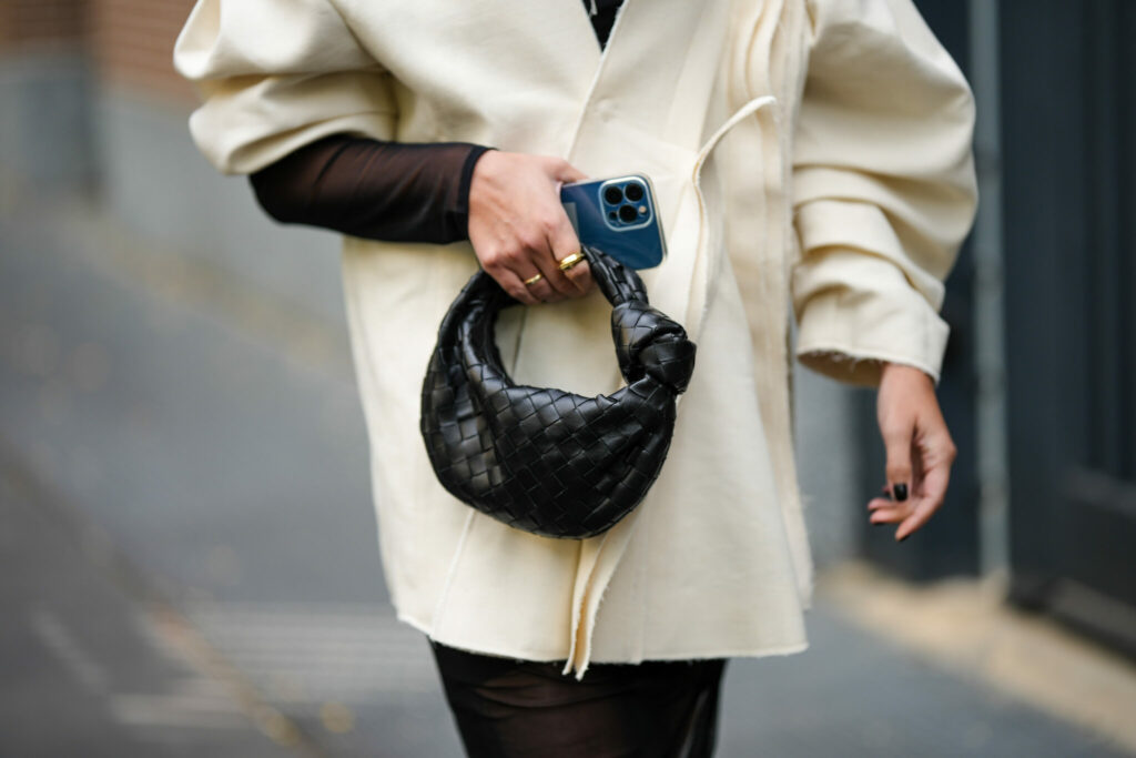 MILAN, ITALY - SEPTEMBER 21: A guest wears a white latte oversized coat, a black asymmetric shoulder / long sleeves / asymmetric midi dress, a black shiny braided leather Jodie handbag from Bottega Veneta, outside Fendi, during the Milan Fashion Week - Womenswear Spring/Summer 2023 on September 21, 2022 in Milan, Italy. (Photo by Edward Berthelot/Getty Images)