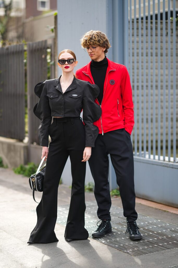 Femeie în total look negru și bărbat în pulover negru cu guler înalt, în pantaloni negri și geacă roșie