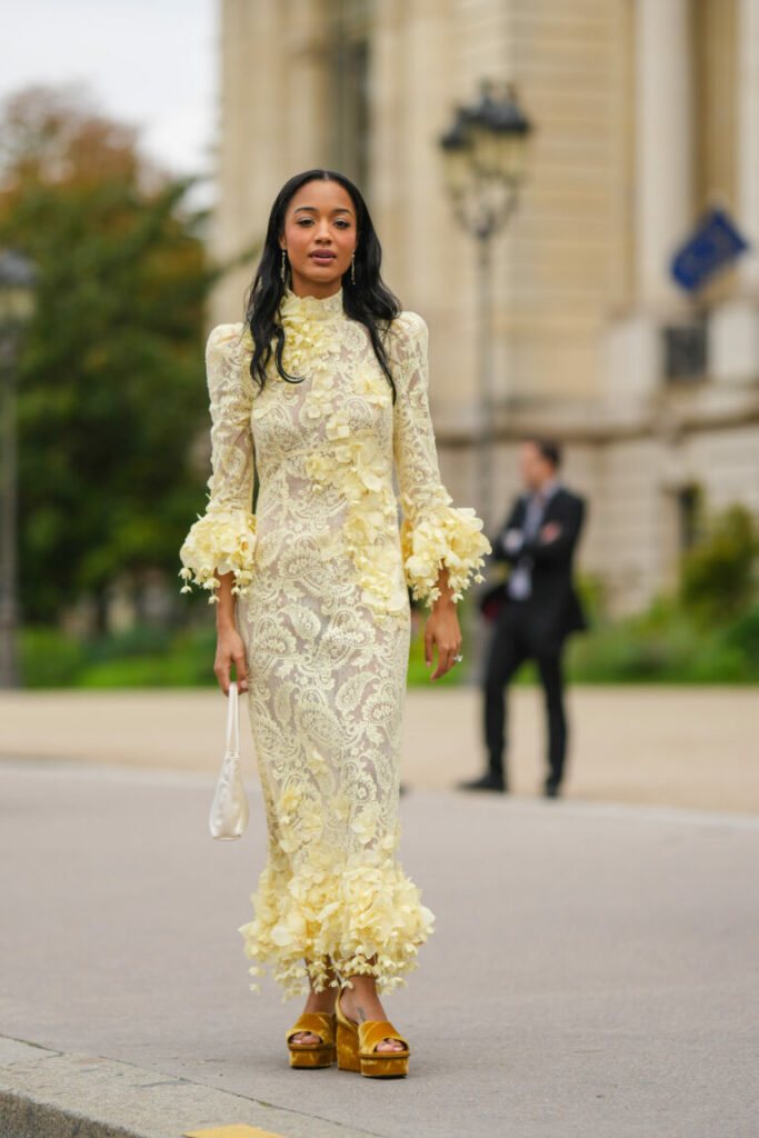 O invitată poartă cercei cu pandantiv auriu, o rochie galben pal cu guler înalt cu model cu imprimeu de dantelă / umeri bufanți / mâneci lungi / rochie lungă, o geantă de mână albă din piele lucioasă, un inel de argint și diamante, sandale cu tocuri bloc din catifea galbenă cu barete , în fața Zimmermann, Paris Fashion Week - Womenswear Spring/Summer 2023