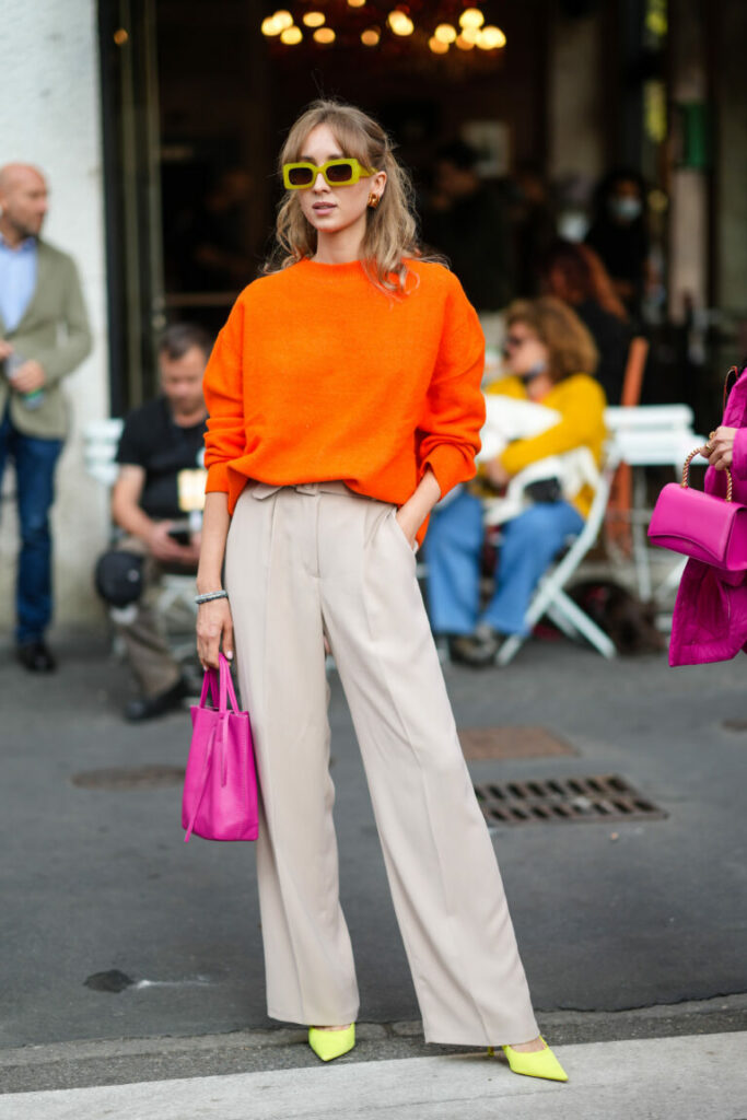o femeie poartă pantaloni eleganți largi, pantofi stiletto, un pulover portocaliu și o geantă roz
